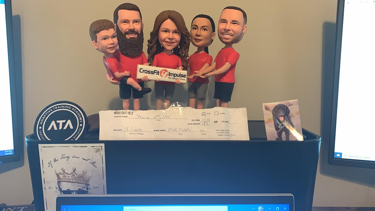 A shelf displaying five bobblehead figurines of people in red shirts, holding a CrossFit sign. Below, there is a workout score sheet, a round ATA logo, a decorative art piece, and a photograph of a black dog.