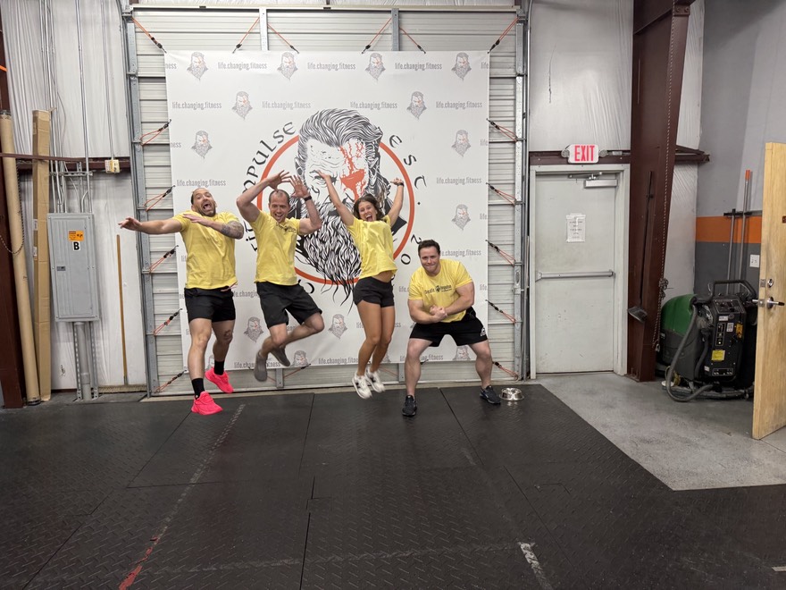 A group of four people wearing yellow shirts jumps and poses energetically in a gym setting with a branding backdrop.