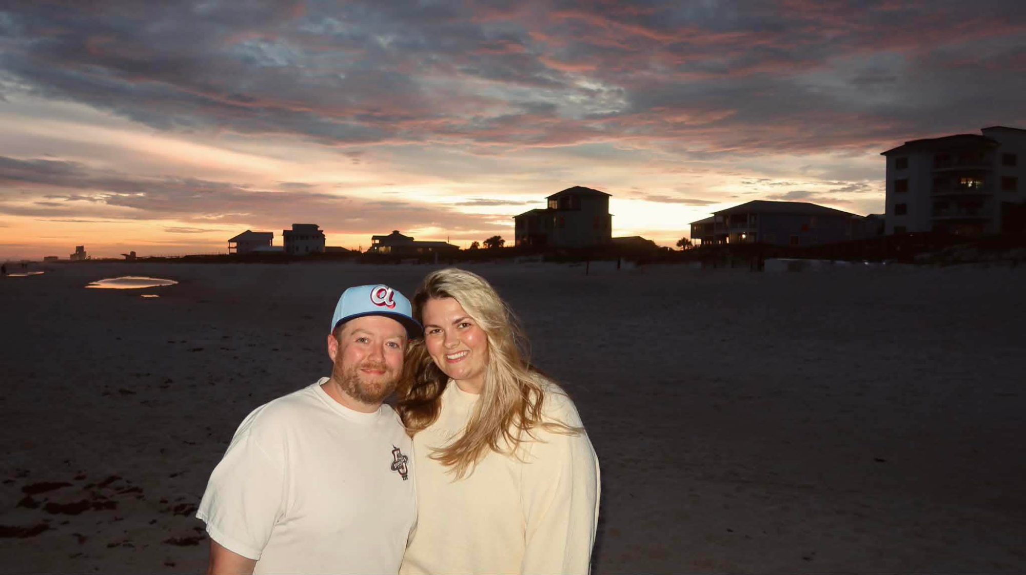 A couple posing together on a beach at sunset, with colorful clouds in the background and beach houses silhouetted against the horizon.