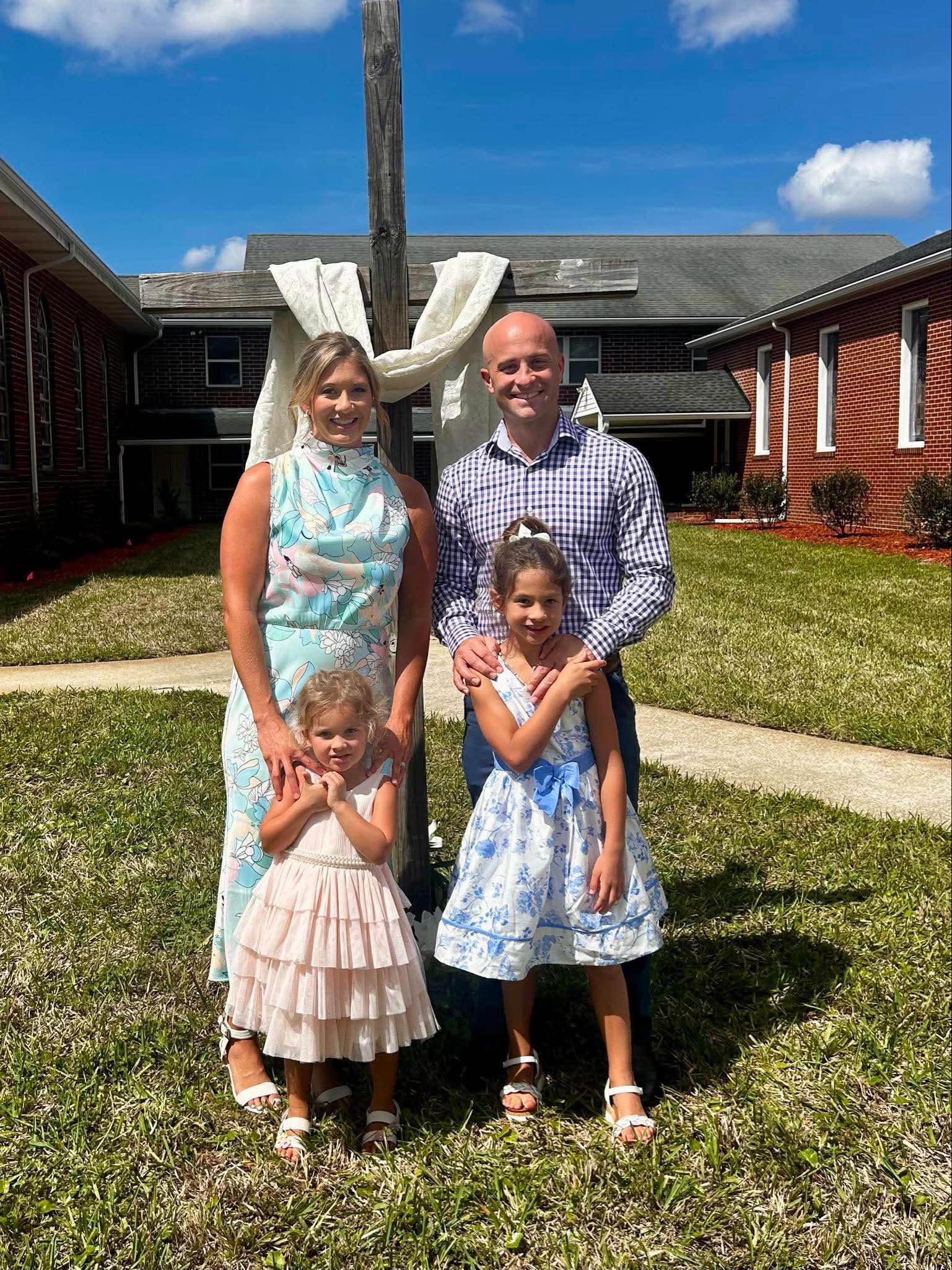 A family posing outdoors in front of a decorative wooden cross, with two young girls and their parents. The girls are wearing light-colored dresses, and the parents are dressed in formal attire. The background shows a sunny day with blue skies and a grassy area.