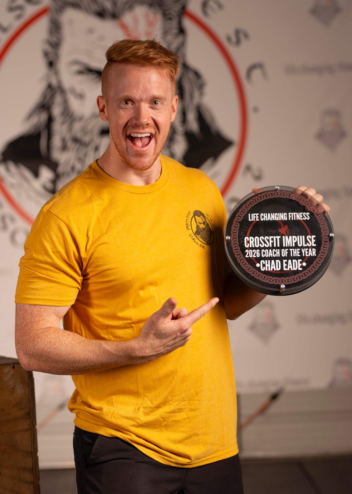 A man with ginger hair wearing a yellow shirt is excitedly holding a plaque that reads 'Life Changing Fitness - Crossfit Impulse 2026 Coach of the Year - Chad Eade'.